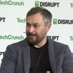A bearded man grins against a TechCrunch-branded backdrop
