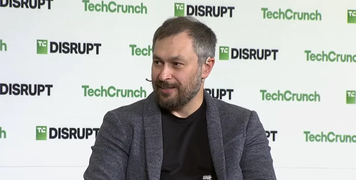 A bearded man grins against a TechCrunch-branded backdrop
