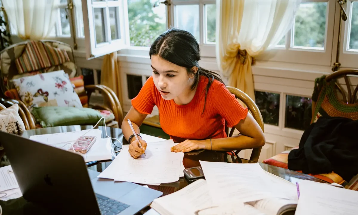 hispanic latina college student works on assignment at home. She is writing something on paper. A laptop is on her desk.