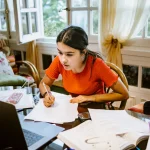 hispanic latina college student works on assignment at home. She is writing something on paper. A laptop is on her desk.