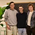 Three smiling men stand in front of a chalkboard