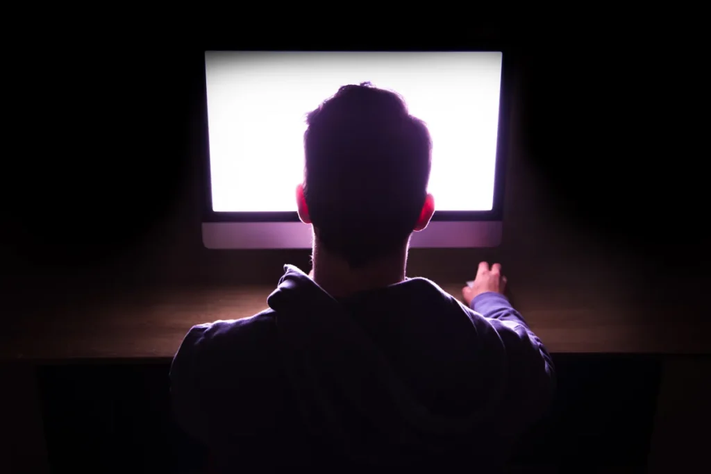 Back view of a man in front of bright computer working in the dark.