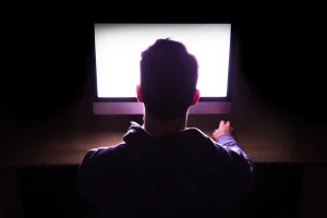 Back view of a man in front of bright computer working in the dark.