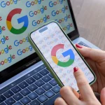 A woman looks at a mobile phone displaying the logo of Google in front of a laptop screen displaying the logo of Google.