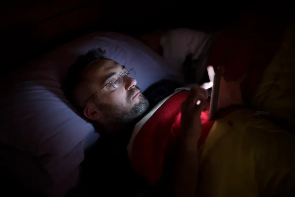 A man in glasses lies in bed, illuminated by the soft glow of his smartphone screen.
