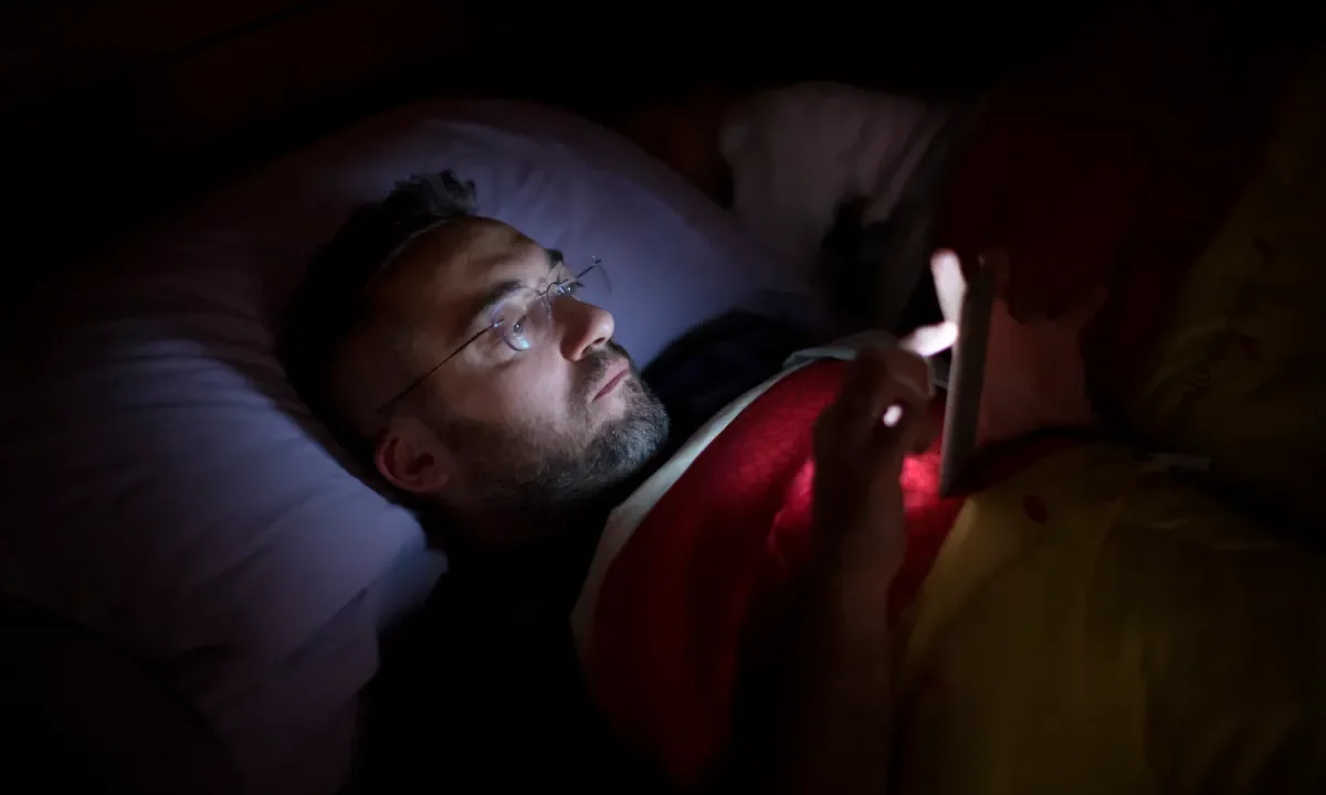 A man in glasses lies in bed, illuminated by the soft glow of his smartphone screen.