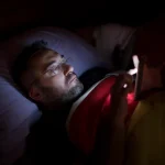 A man in glasses lies in bed, illuminated by the soft glow of his smartphone screen.