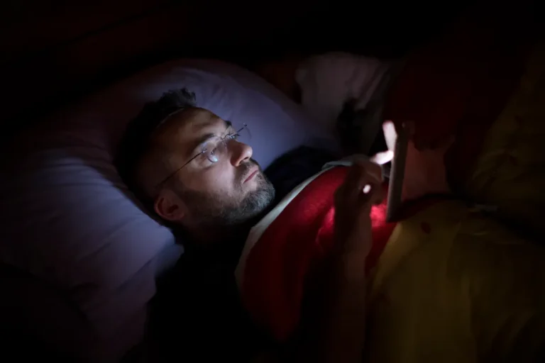 A man in glasses lies in bed, illuminated by the soft glow of his smartphone screen.