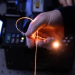 A hand holds a single strand of fiber in a darkened room, with light pouring from its tip.