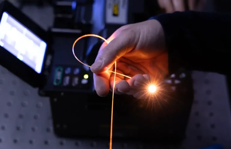 A hand holds a single strand of fiber in a darkened room, with light pouring from its tip.