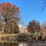 A wooded lake, with a stately Georgian brick building visible in the distance.