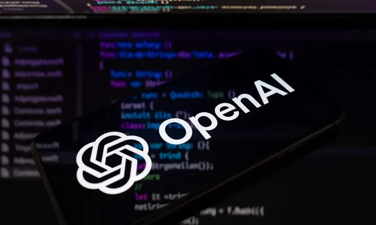 The OpenAI logo is displayed on a smartphone screen placed on a reflective surface onto which lines of computer code.