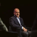A man in a business suit sits on a conference stage